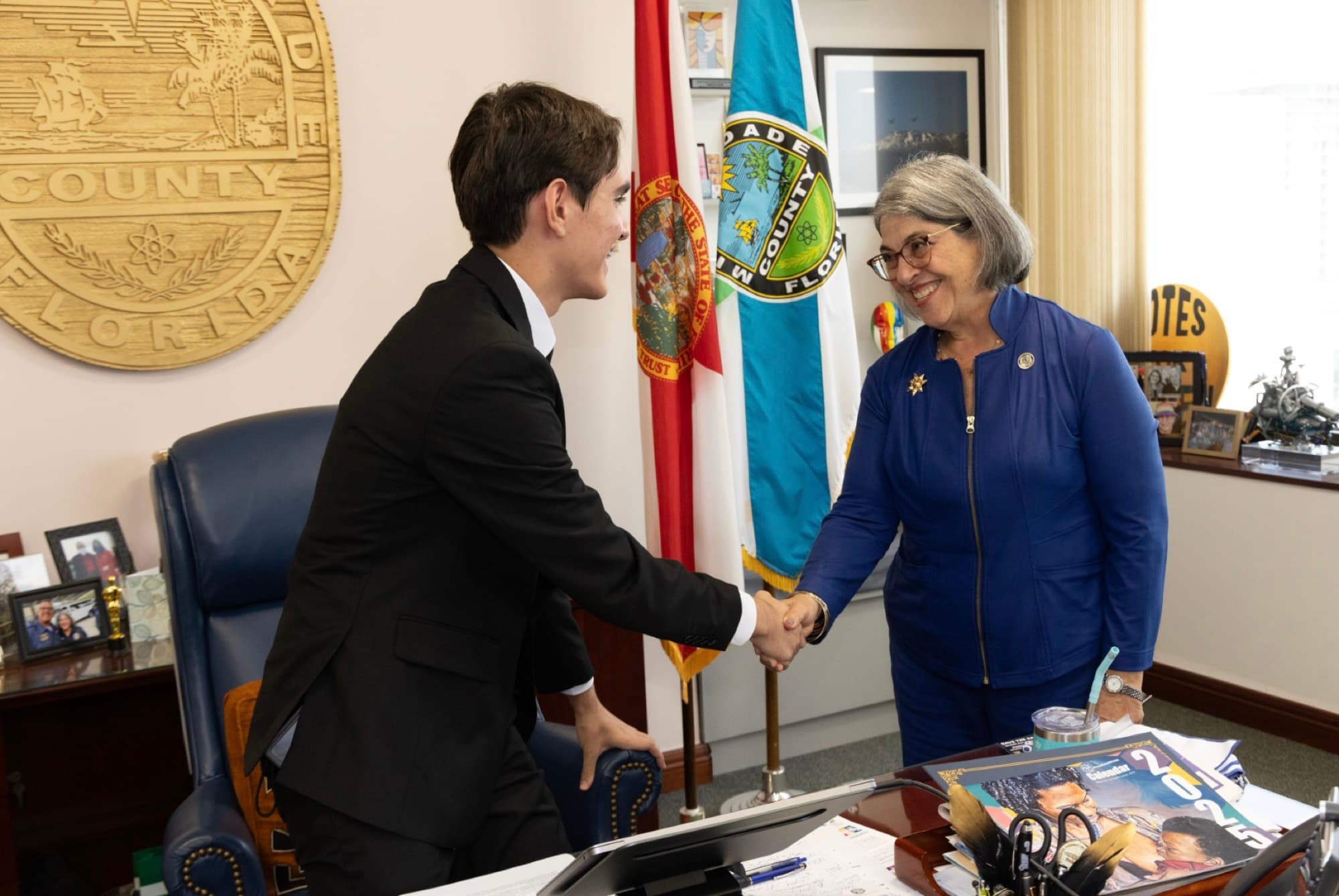 A young man in a dark suit shakes hands with an official across a desk in a Miami-Dade County office.