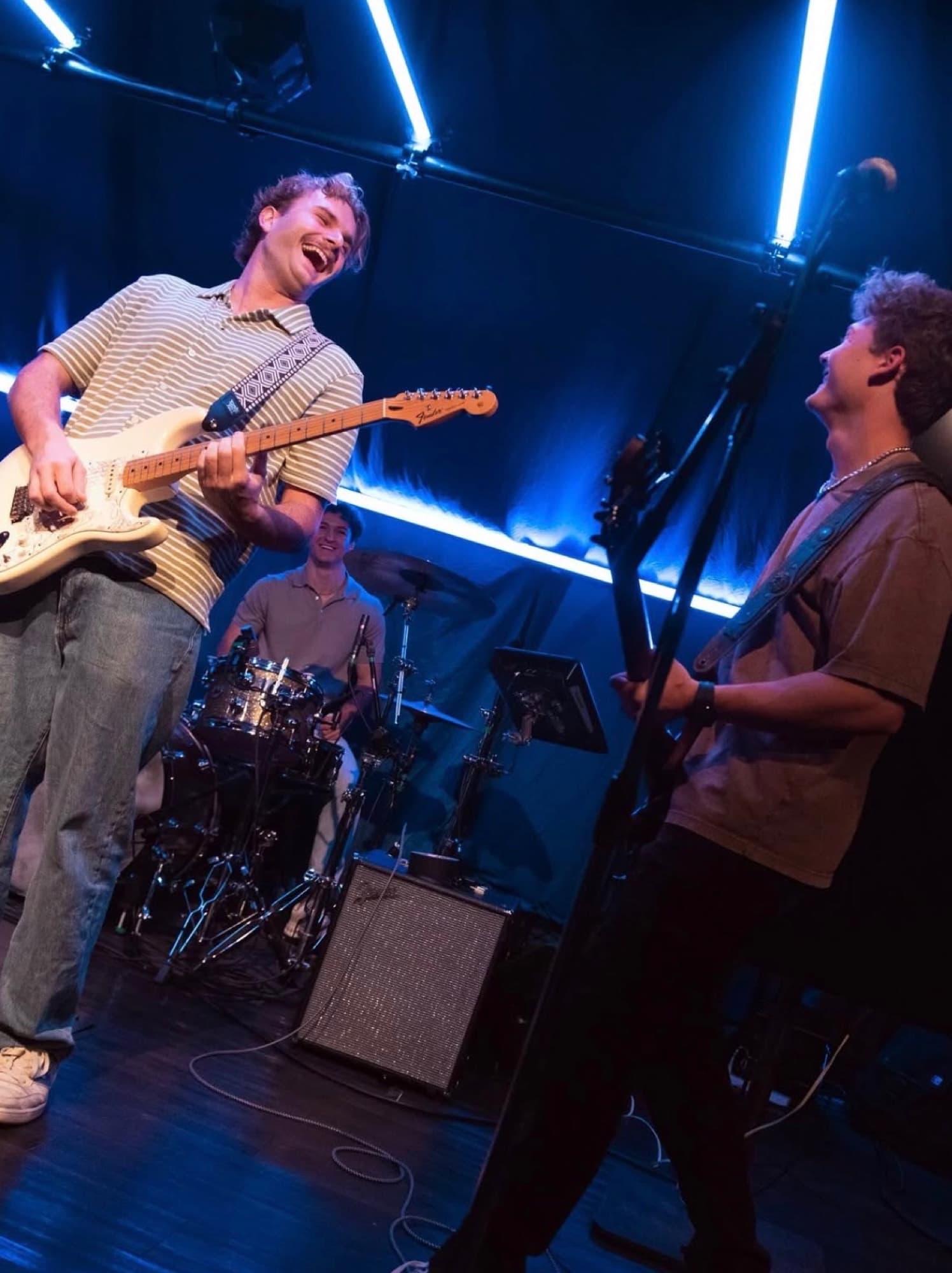 A band laughing together mid-song on a small stage lit by blue tube lights.
