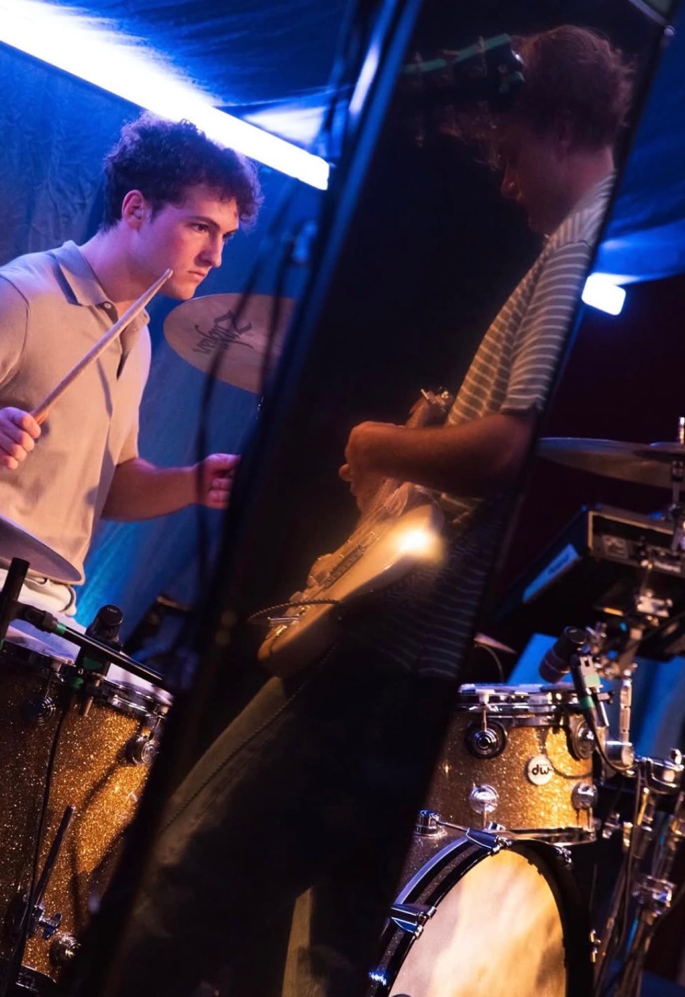 A drummer and a guitarist seen through a narrow gap backstage, gold sparkle drum kit catching the light.